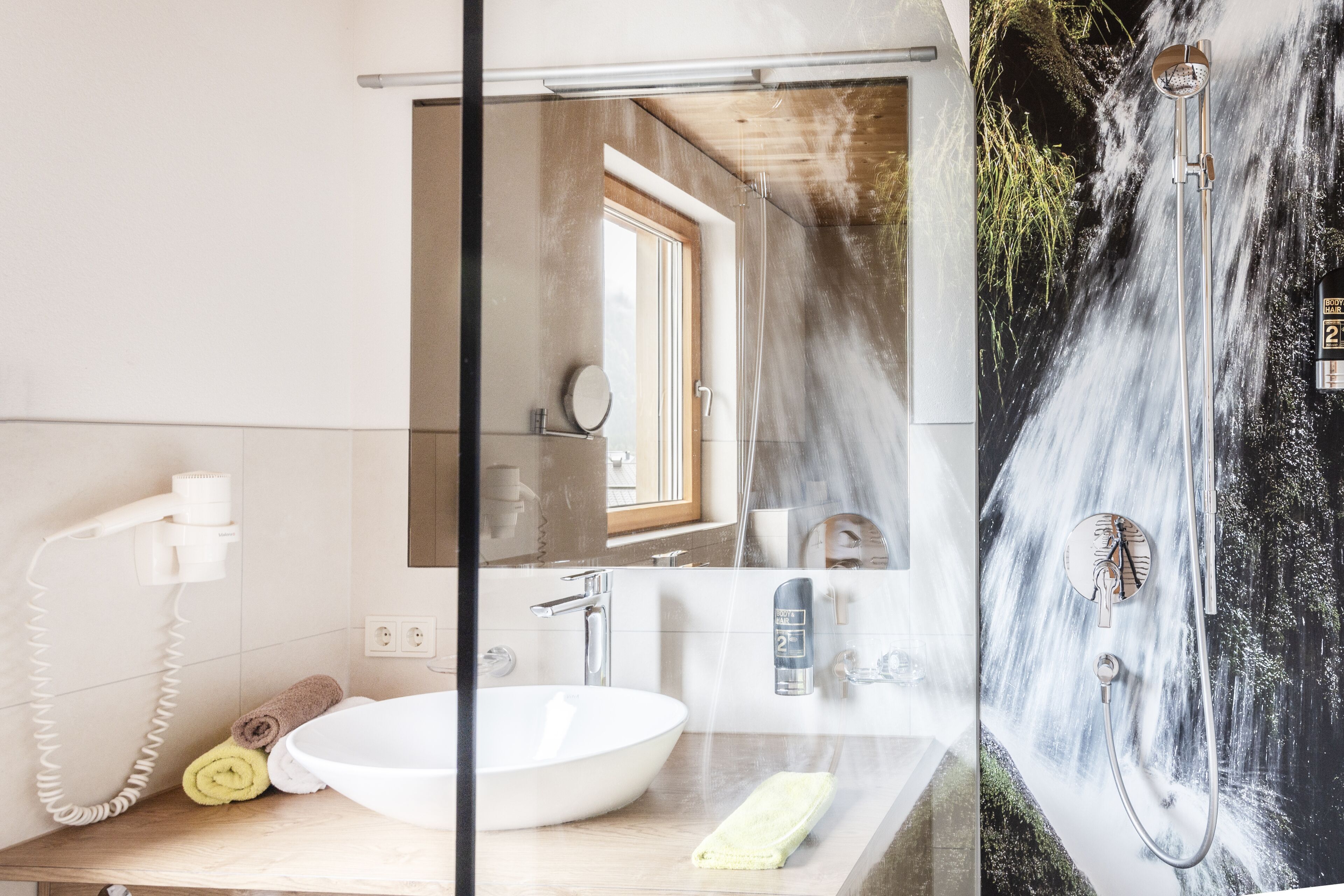 A bathroom with a large sink, a mirror, a hairdryer, and a shower with a waterfall mural.