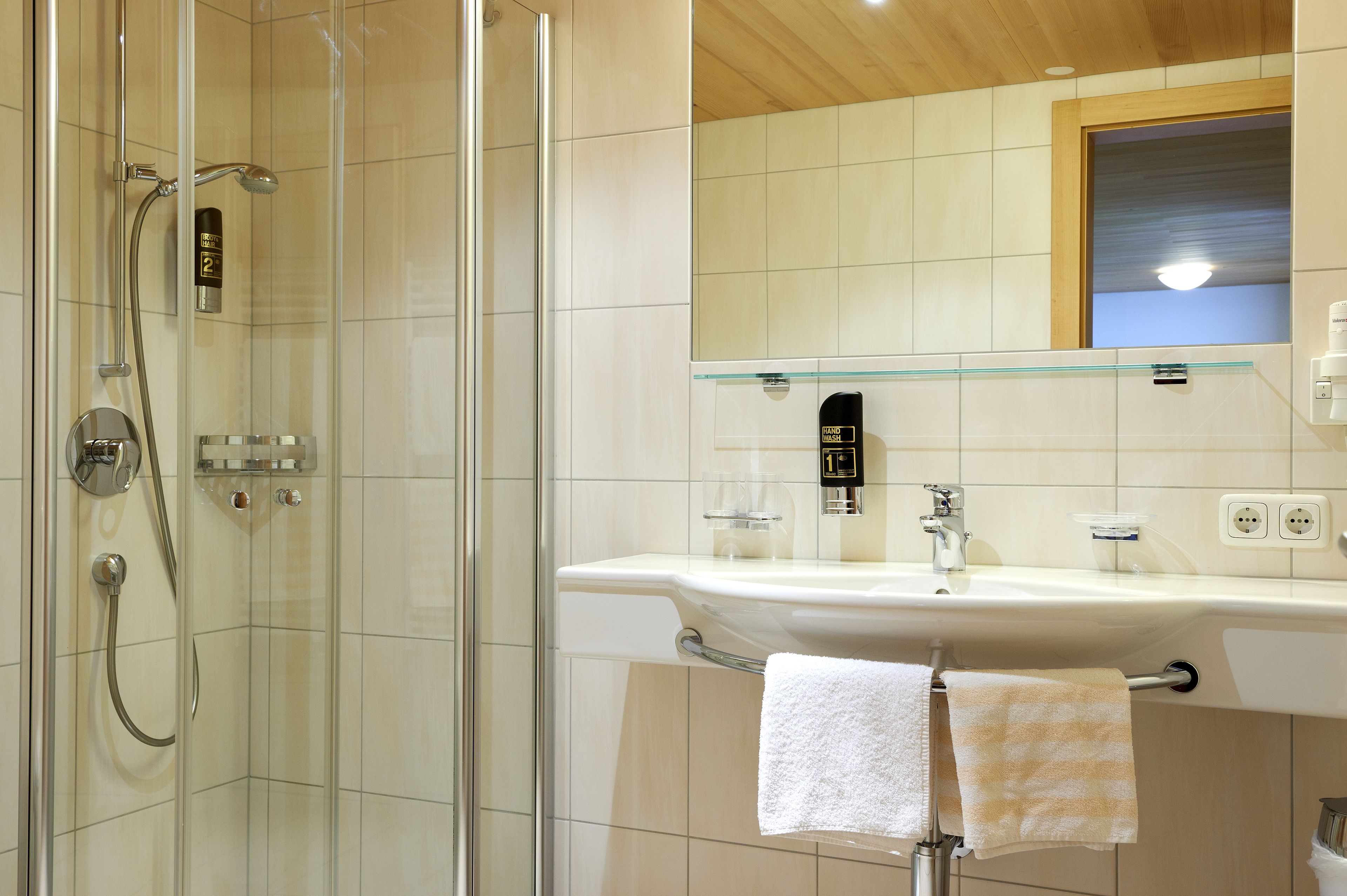 A modern bathroom with a glass-enclosed shower, a white sink with a glass shelf, and a wooden ceiling.