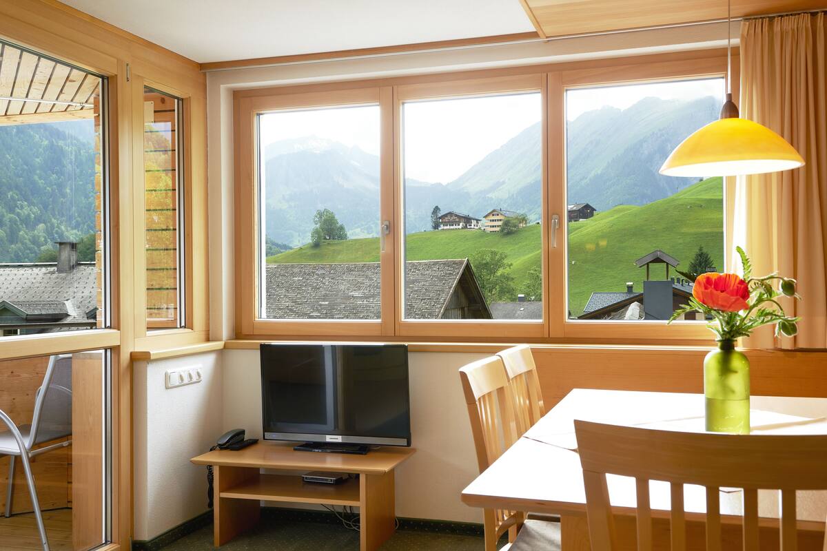 Una habitación con una televisión sobre un soporte de madera, una mesa de comedor con sillas, un jarrón con flores y ventanas amplias con vista a la montaña.