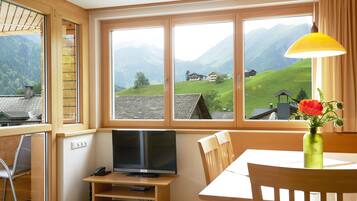 A room with a TV on a wooden stand, a dining table with chairs, a vase with flowers, and large windows with a mountain view.
