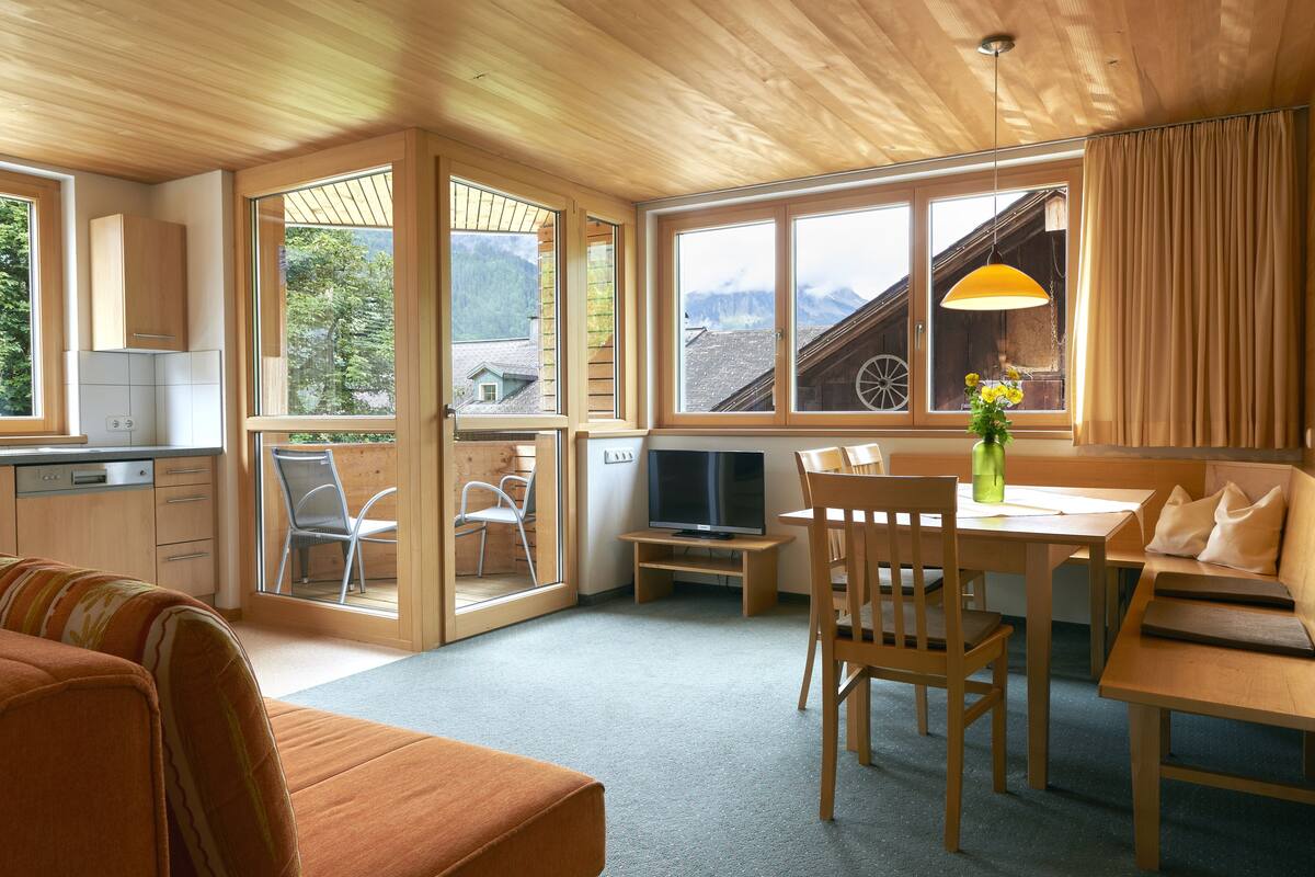 Interior acogedor de una cabaña con paredes y suelos de madera, zona de comedor con mesa y sillas, sofá, televisor y ventanas amplias con vistas a la montaña.