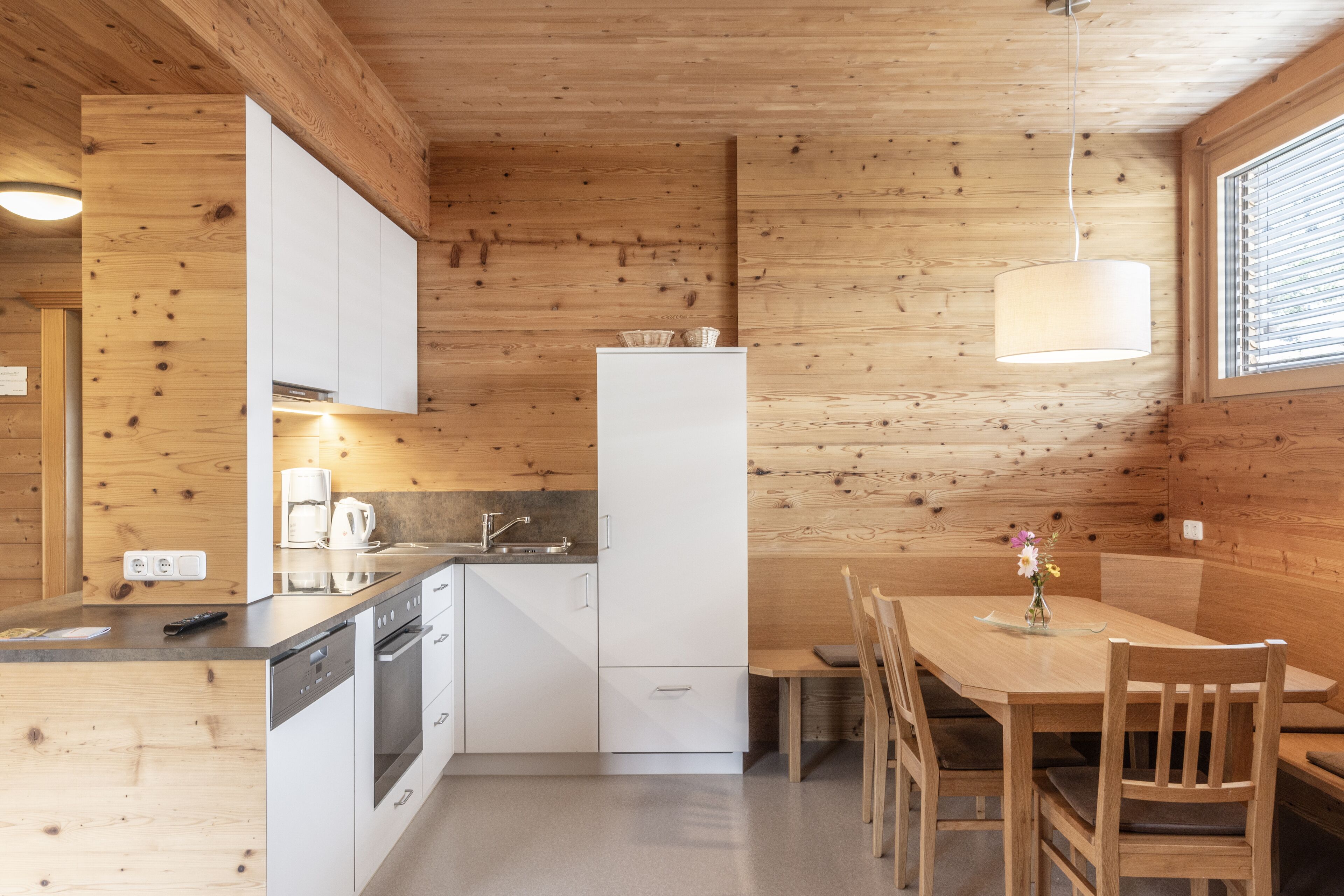 A compact kitchen with white cabinets, a built-in oven, and a refrigerator. There is a dining table with chairs and a hanging light fixture.