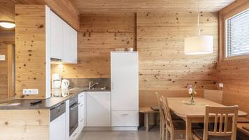 A compact kitchen with white cabinets, a built-in oven, and a refrigerator. There is a dining table with chairs and a hanging light fixture.