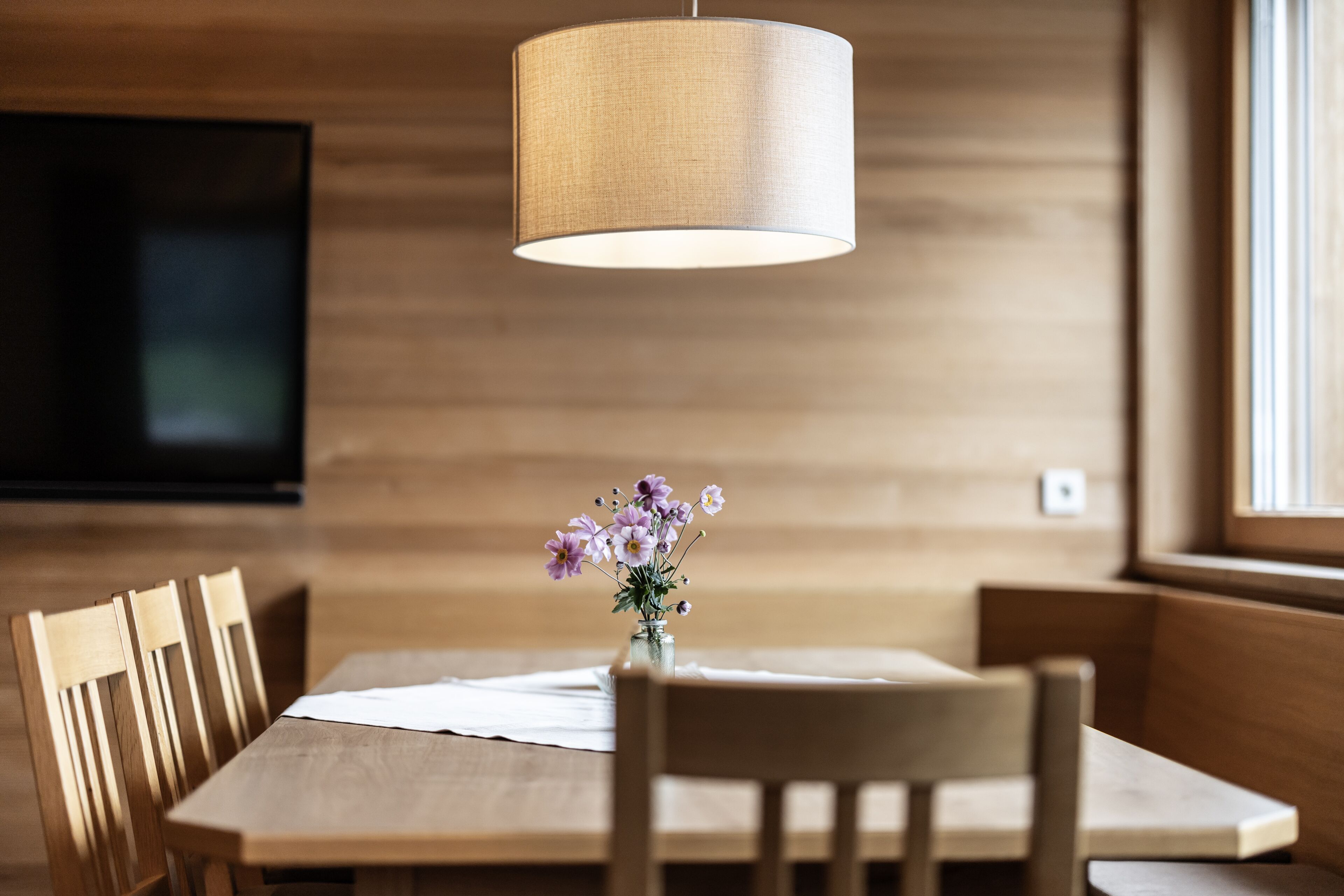A dining area with a wooden table, chairs, a vase with flowers, and a hanging lamp.