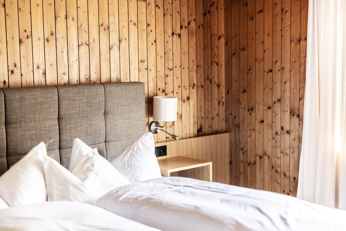 Una cama bien hecha con sábanas y almohadones blancos, cabecera de madera, mesita de noche con lámpara y pared de madera con diseño de tablones verticales.