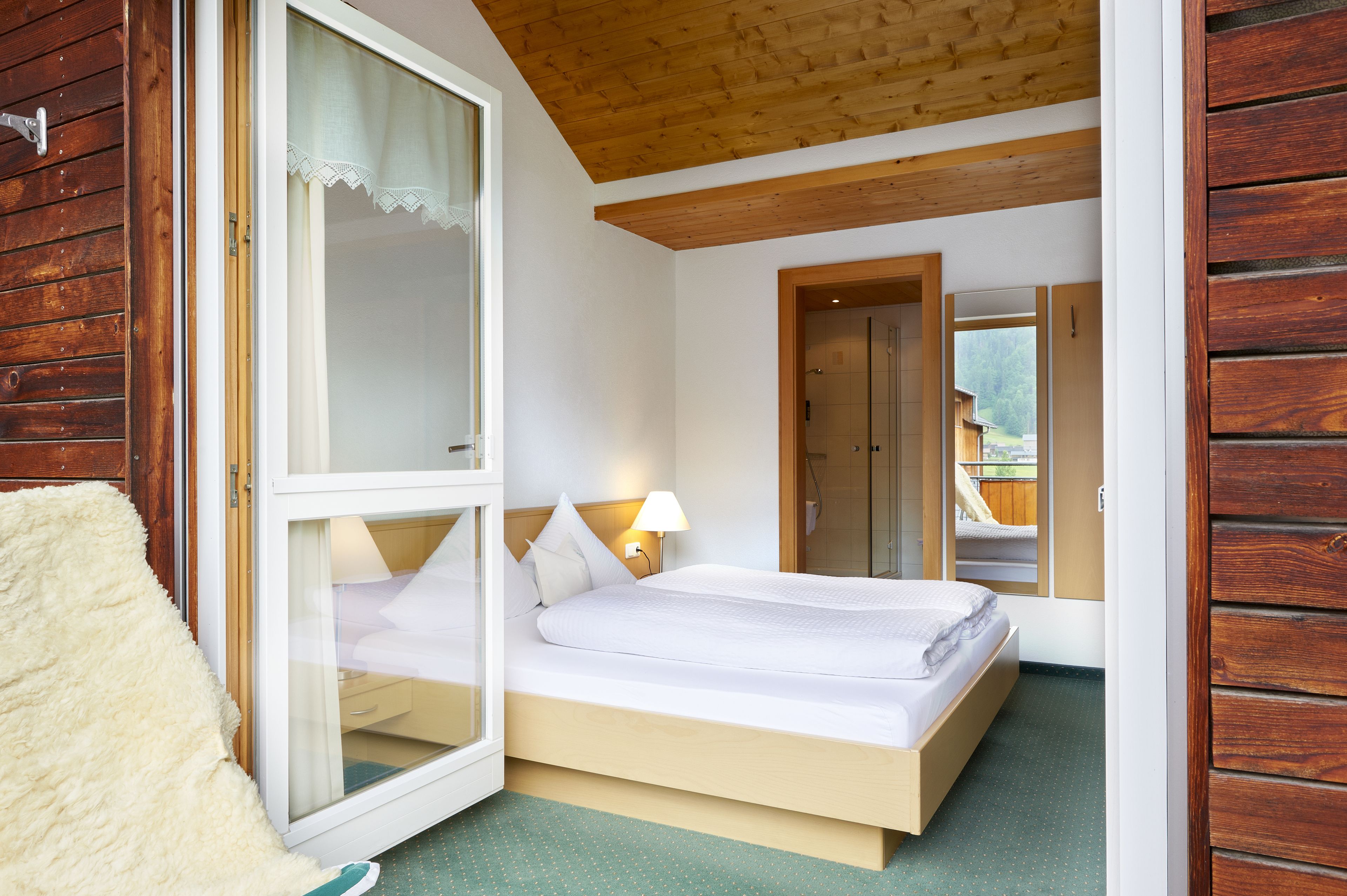 Ein Schlafzimmer mit Holzboden, einem Bett mit weißer Bettwäsche, einem Nachttisch mit Lampe und Blick auf eine Holzhütte durch ein Fenster.