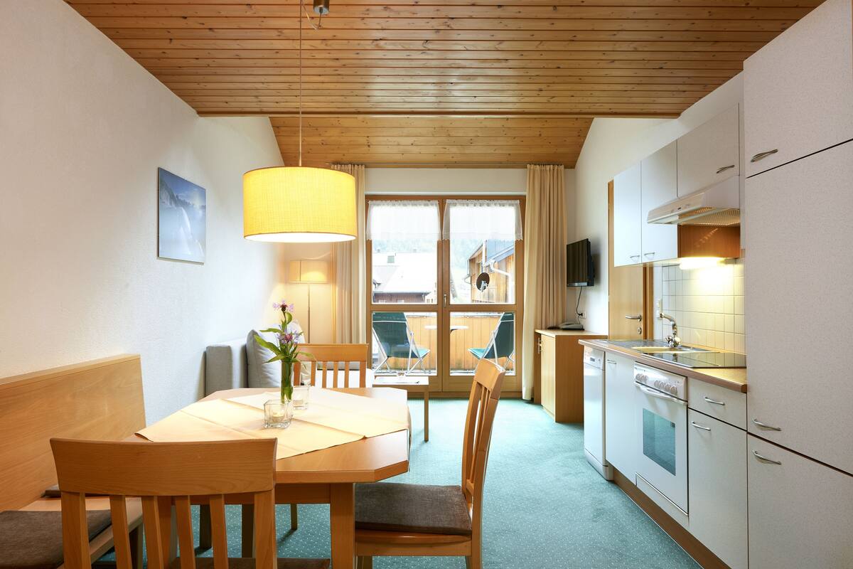 Una cocina funcional con armarios de madera, una mesa de comedor con sillas, una ventana con cortinas y una lámpara colgante.
