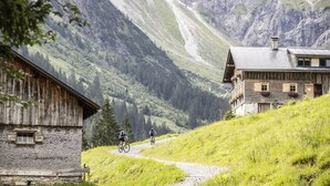 Une cabane de montagne, accessible par un sentier de terre, entourée d’une végétation luxuriante et de montagnes.