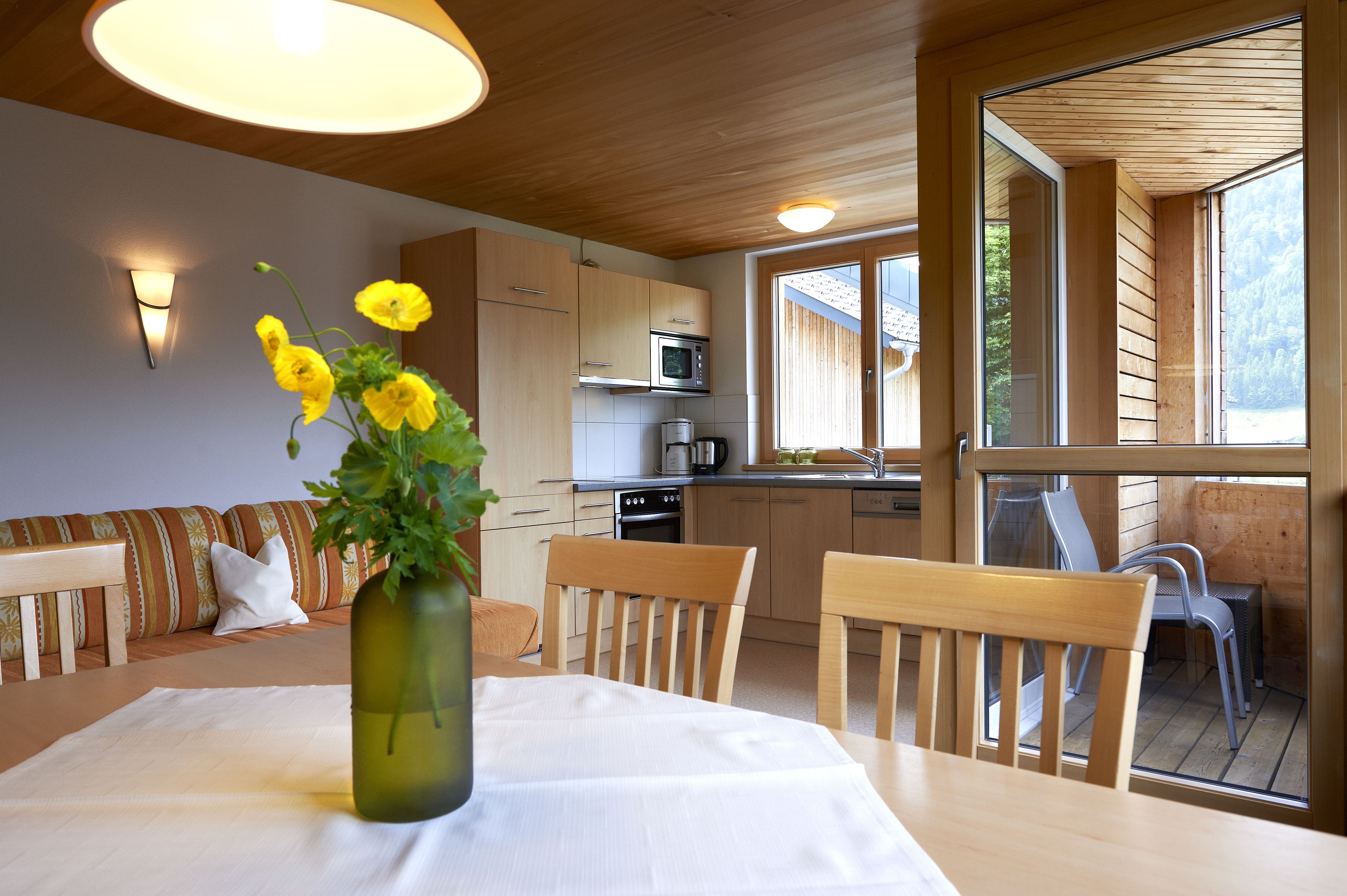 A wooden interior with a dining table, chairs, a vase with flowers, and a kitchen area with a microwave and stove.