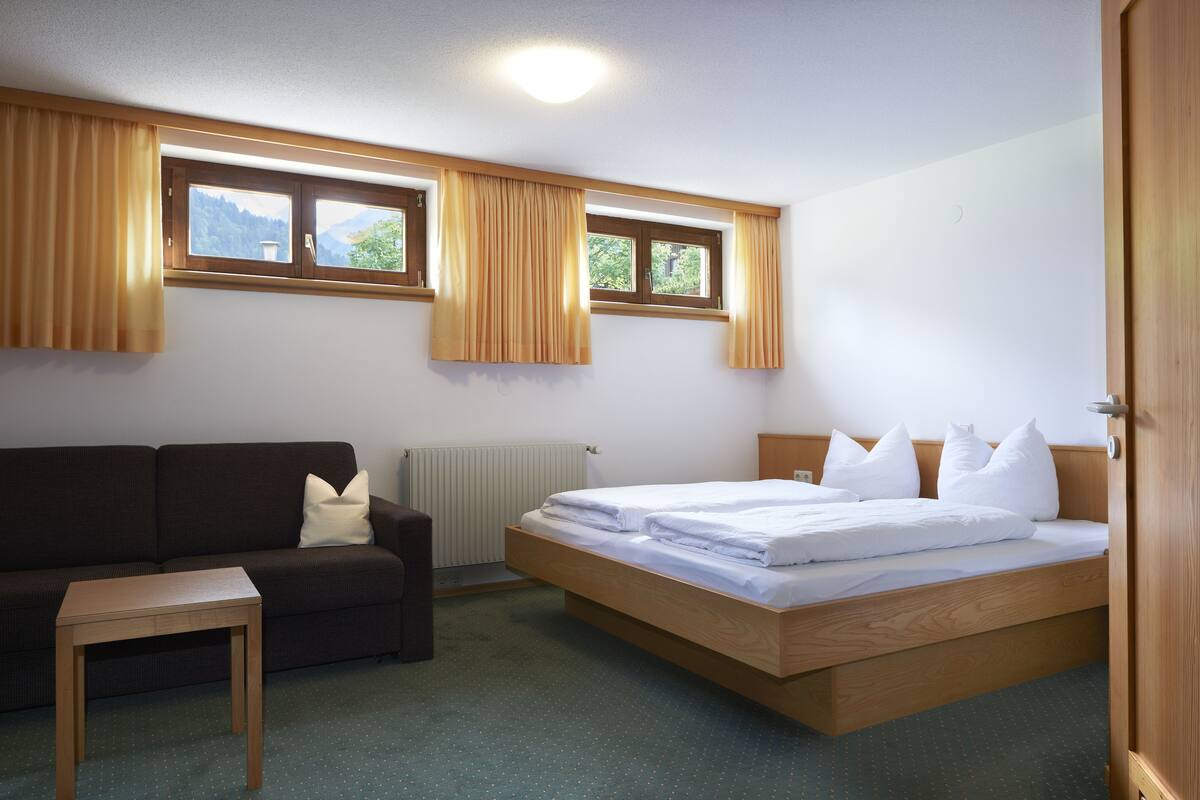 Una habitación con cama, sofá, una mesita y dos ventanas con cortinas.