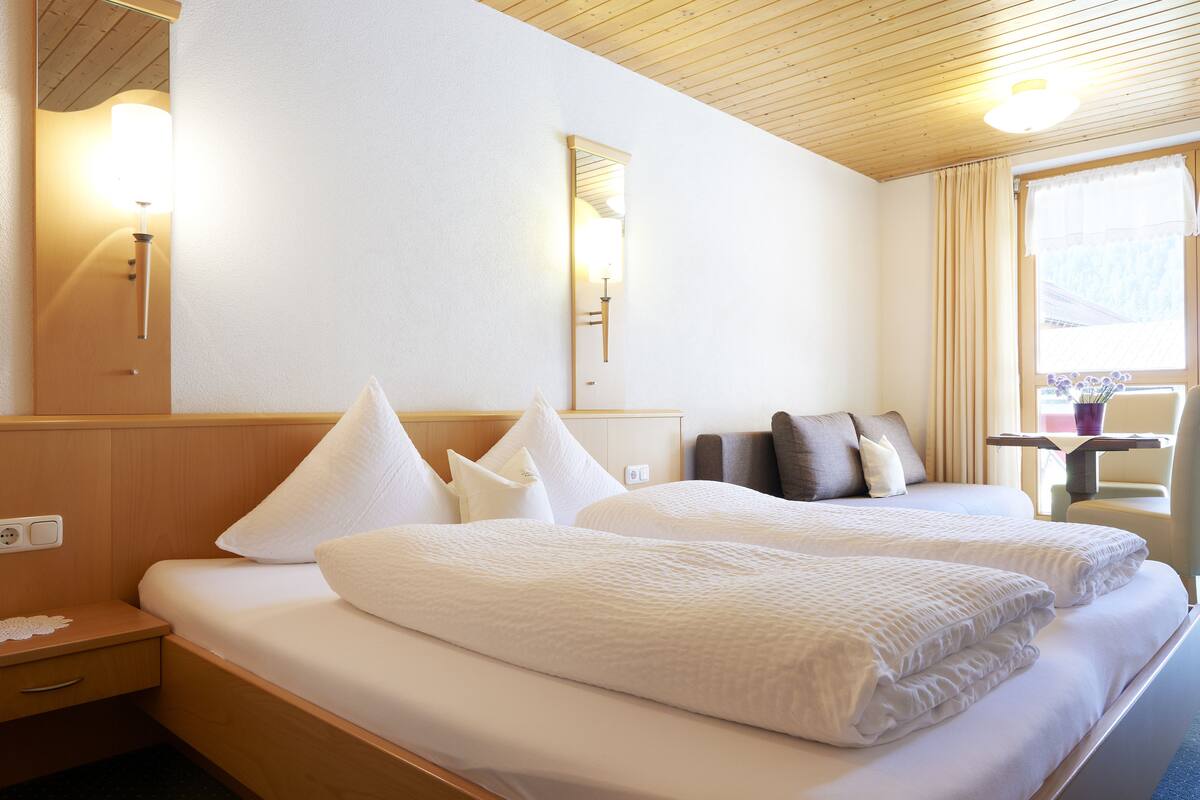 Habitación de hotel con dos camas, cabecera de madera, mesita de noche con lámpara, ventana con cortinas y vistas al exterior.