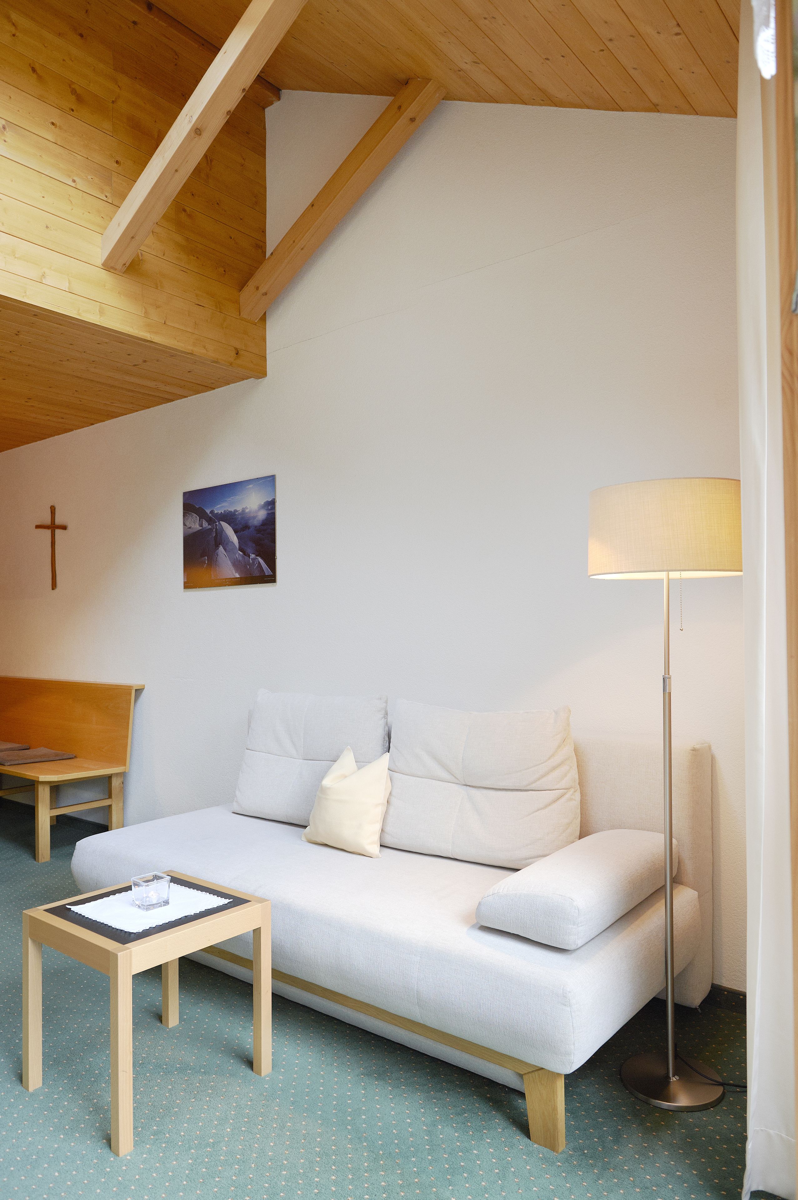 A cozy room with a wooden ceiling, a white sofa, a small wooden table, a floor lamp, and a cross on the wall.