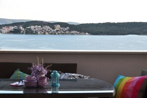 A balcony with a table set for two, overlooking a coastal town with buildings and hills in the background.