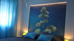 Hotellihuone, jossa on suuri sänky, kaksi sinistä tyynyä, yöpöytä lampun kanssa ja seinällä oleva maalaus, jossa on kuvattuna puu.