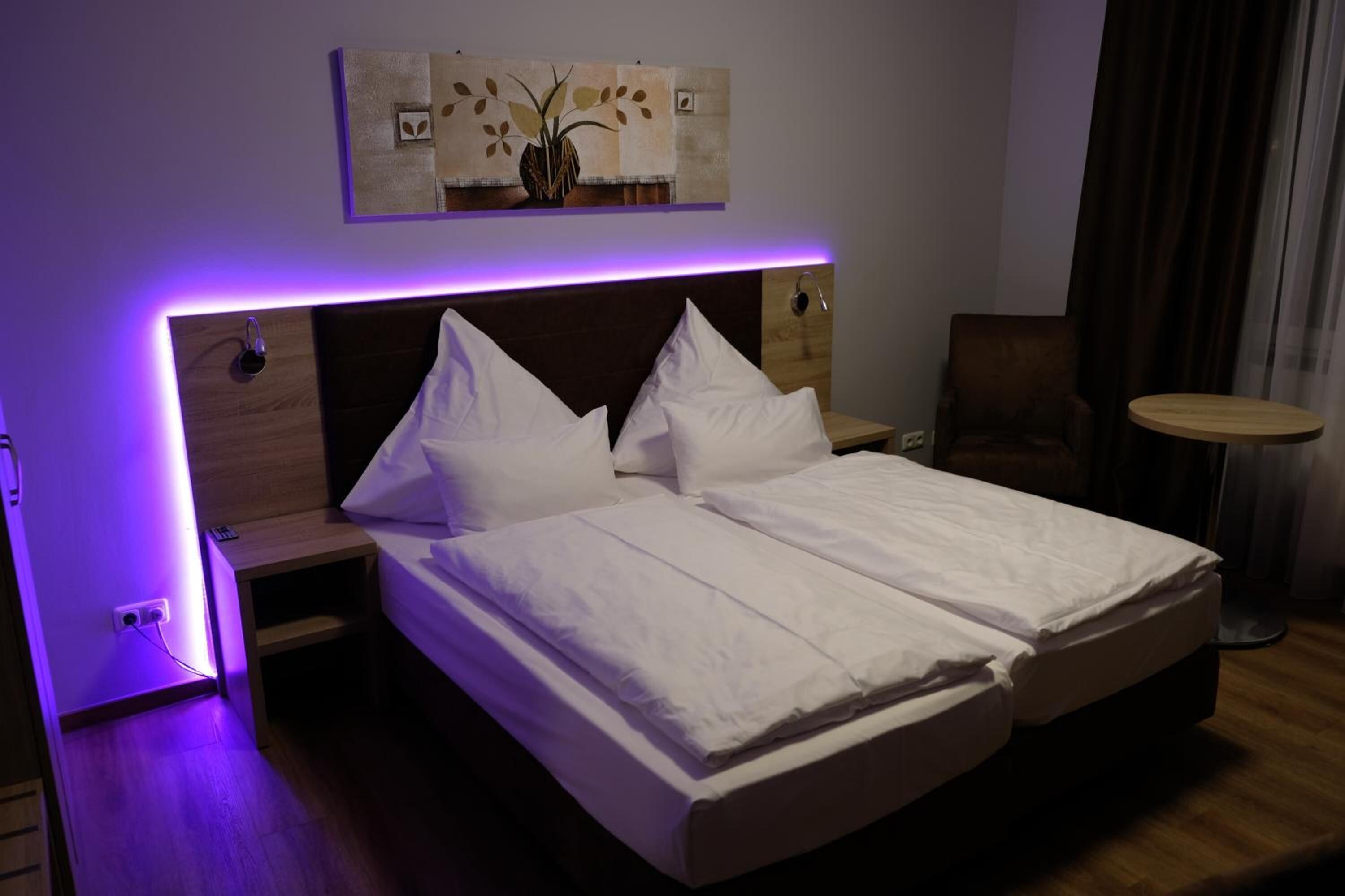 A double bed with white linens, a bedside table with a lamp, a small round table, and a painting above the bed.