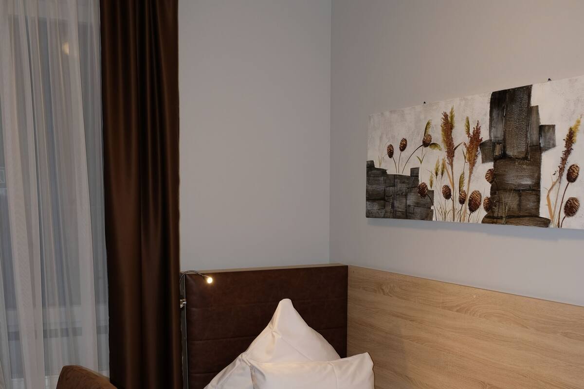 Una cama individual con sábanas blancas, cabecera de madera, una silla marrón y un cuadro en la pared.
