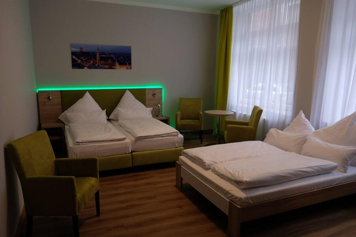 Habitación de hotel con cama doble, dos camas individuales, un sillón verde, una mesita redonda y vistas a la ciudad.