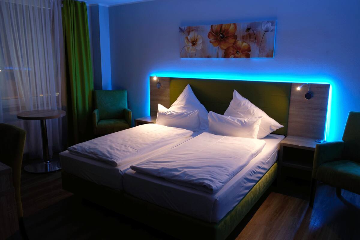 Una cama doble con ropa blanca impecable, cabecera verde y una tira de luz ambiental azul. A la izquierda, una pequeña mesa redonda y una silla verde; a la derecha, un sillón verde.