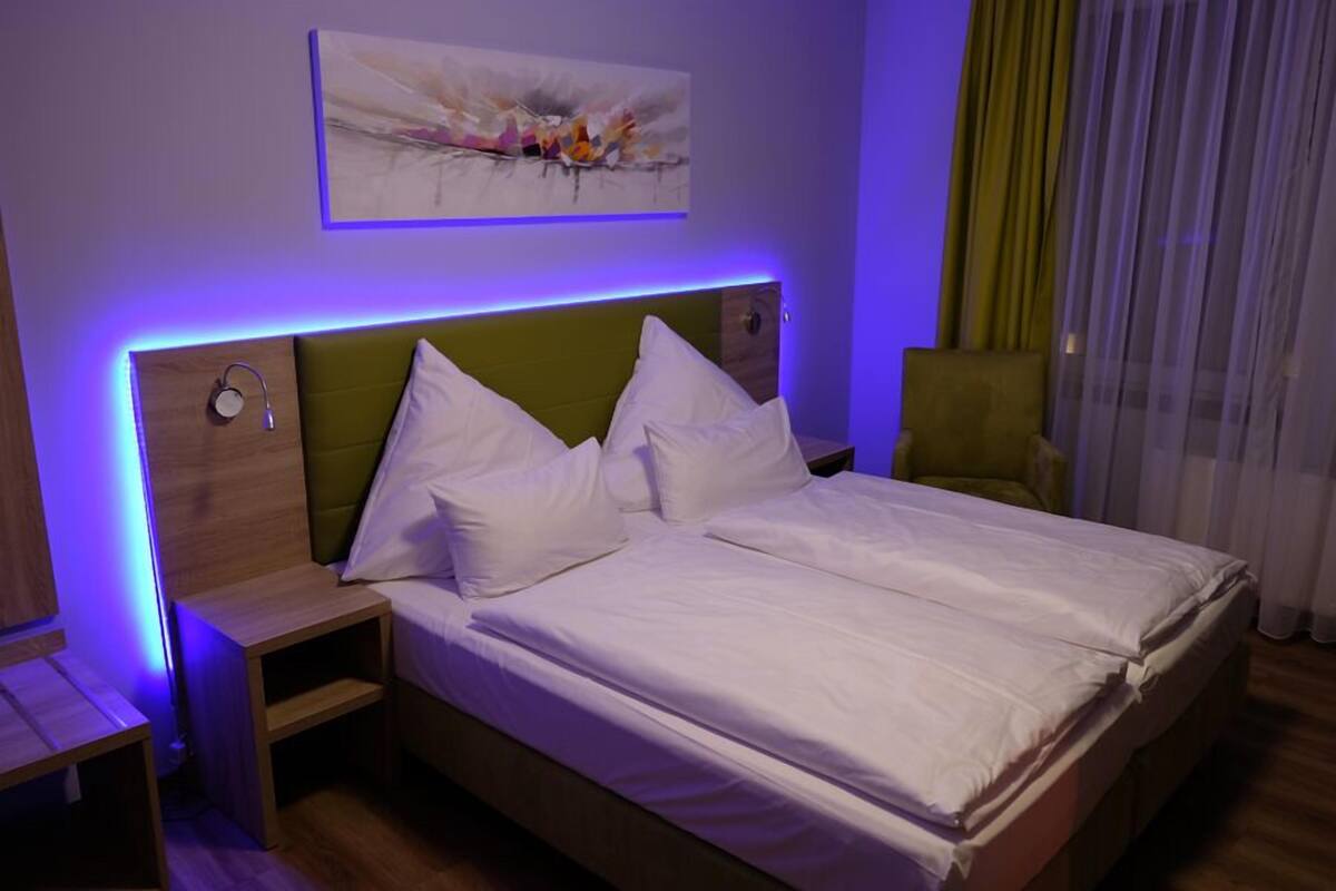 Una cama bien hecha con sábanas y almohadas blancas, un cabecero tapizado en verde y una mesita de noche con una lámpara.
