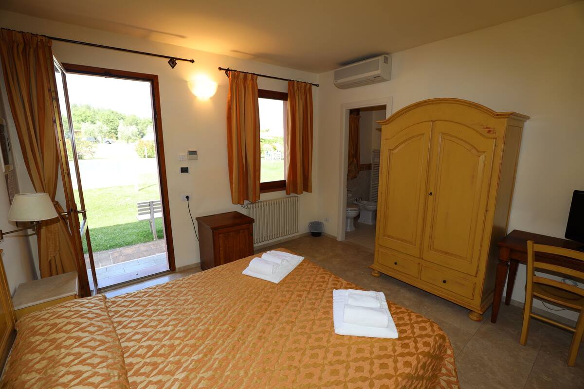 Ein Hotelzimmer mit einem Bett, einem Kleiderschrank, einem Schreibtisch und einem Stuhl. Ein Fenster mit Vorhängen bietet einen Blick ins Freie.