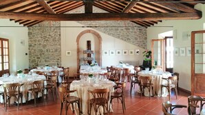 Un vaste espace dédié à la salle à manger, avec plusieurs tables rondes disposées pour les repas, entourées de chaises en bois. La pièce est agrémentée d’un mur en pierre et d’un plafond en bois avec des poutres apparentes.