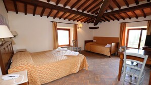 Une chambre d’hôtel avec un grand lit, un plus petit lit, un bureau et une chaise. Il y a deux fenêtres avec des rideaux, une lampe fixée au mur et un plafond en bois.