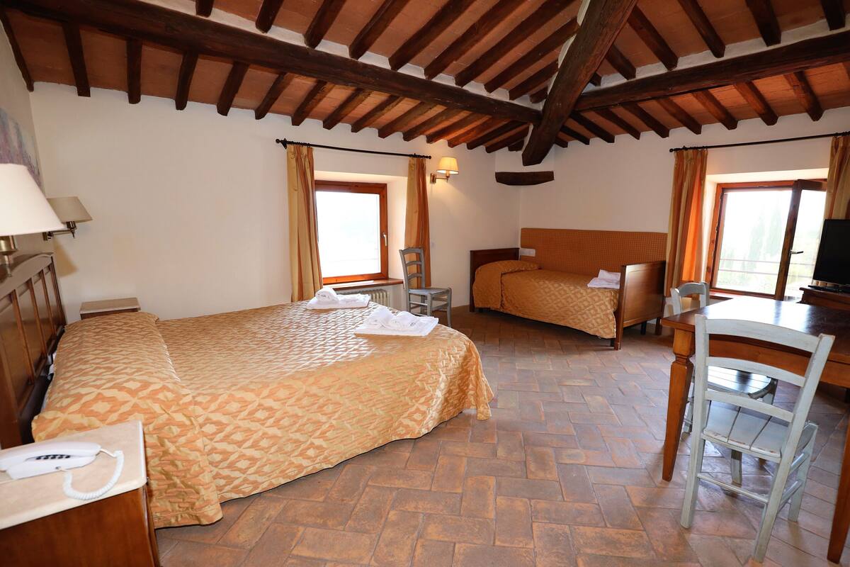 Ein Hotelzimmer mit einem großen Bett, einem Einzelbett, einem Schreibtisch und einem Stuhl. Zwei Fenster mit Vorhängen, eine Wandlampe und eine Holzbalkendecke sind ebenfalls vorhanden.