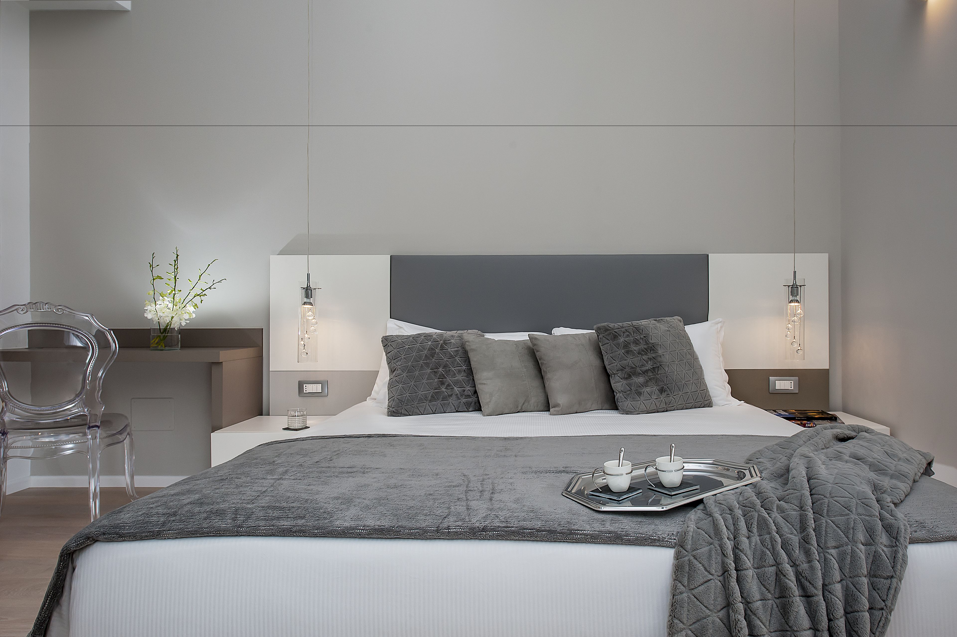 Un dormitorio moderno con una cama grande, ropa de cama gris, una mesita de noche con lámpara y una silla transparente.