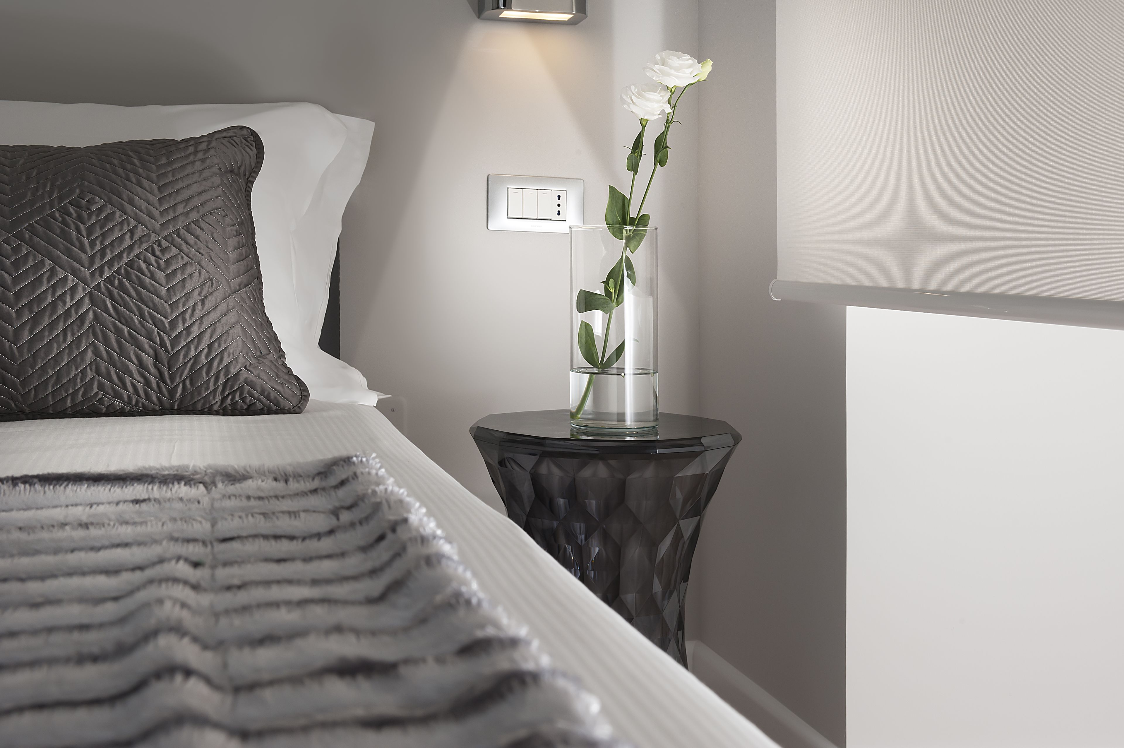 A neatly made bed with a quilted pillow, a bedside table with a glass vase containing a white flower, and a wall-mounted light.