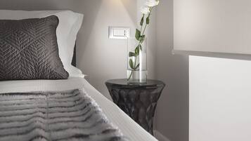 A neatly made bed with a quilted pillow, a bedside table with a glass vase containing a white flower, and a wall-mounted light.