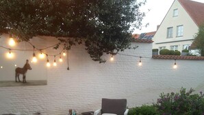 A wooden deck with white furniture, string lights, and a wall-mounted donkey sculpture. - B&B Les Invités (Bruges)