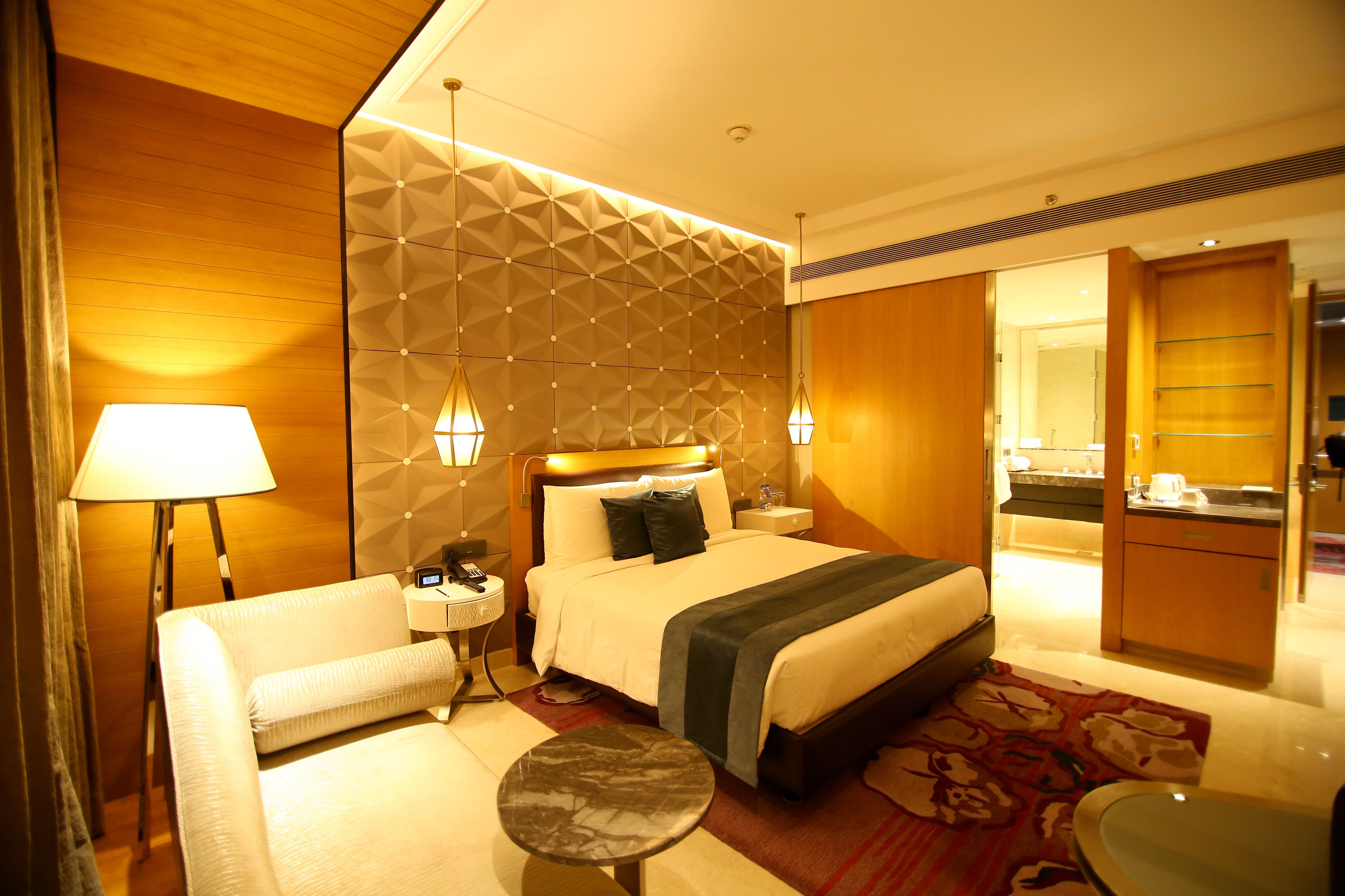 A hotel room with a bed, bedside tables, a lamp, a sofa, a small round table, and a bathroom visible through an open door.