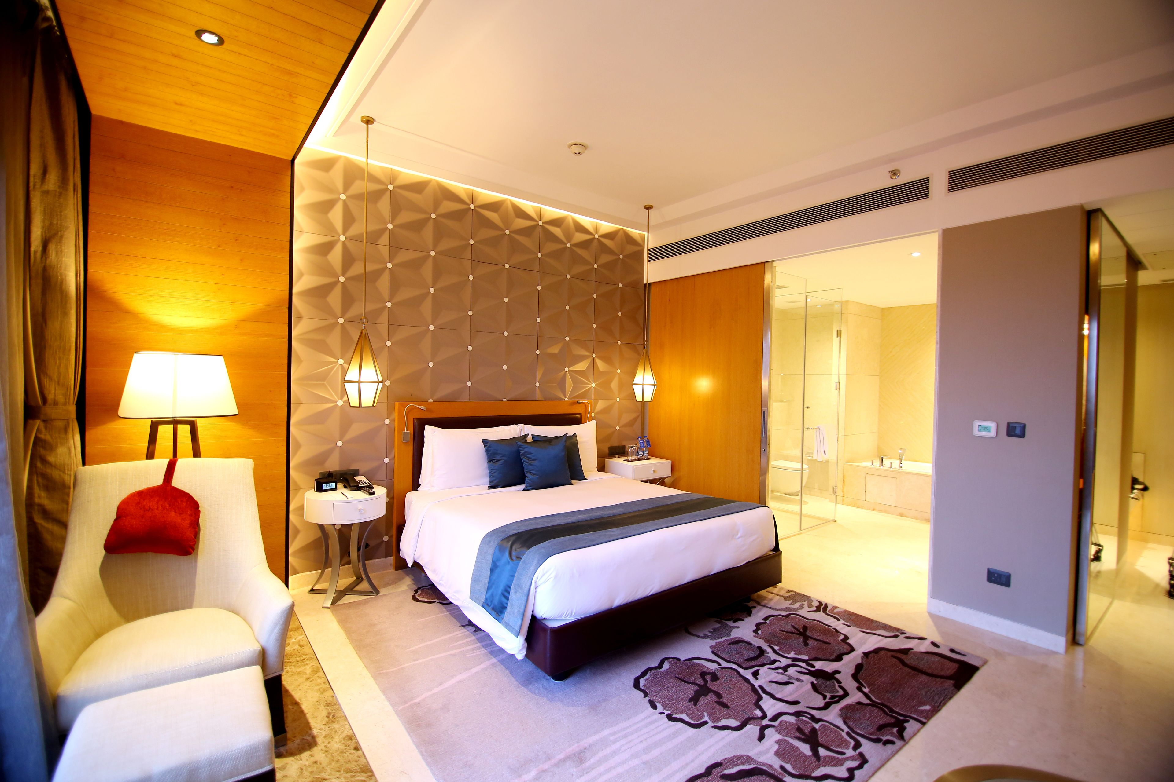 A modern hotel room with a large bed, a sitting area with a chair, a bedside table, and a lamp.