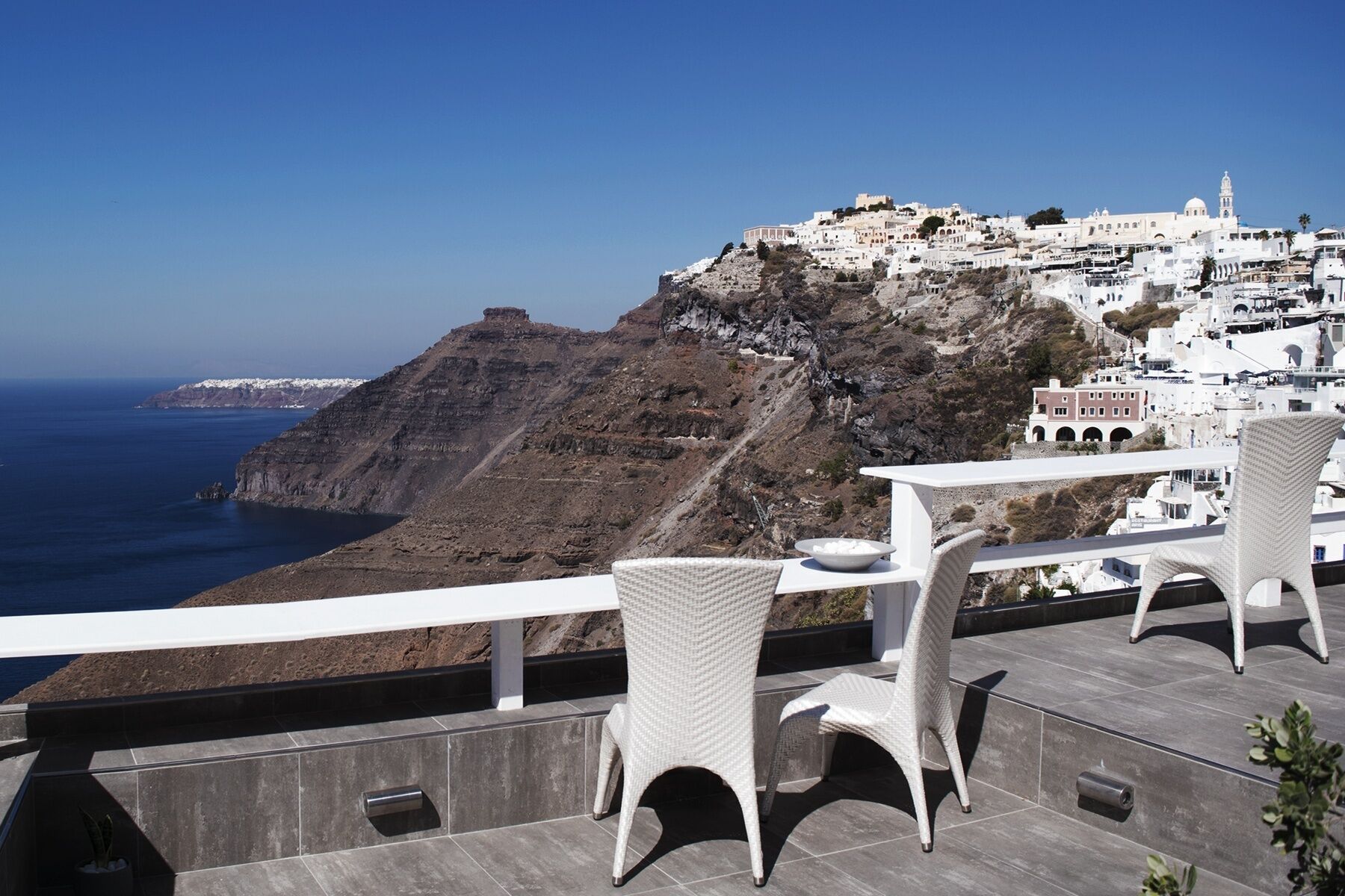 A terrace with white wicker chairs and a table, overlooking a coastal town with white buildings and a cliff.