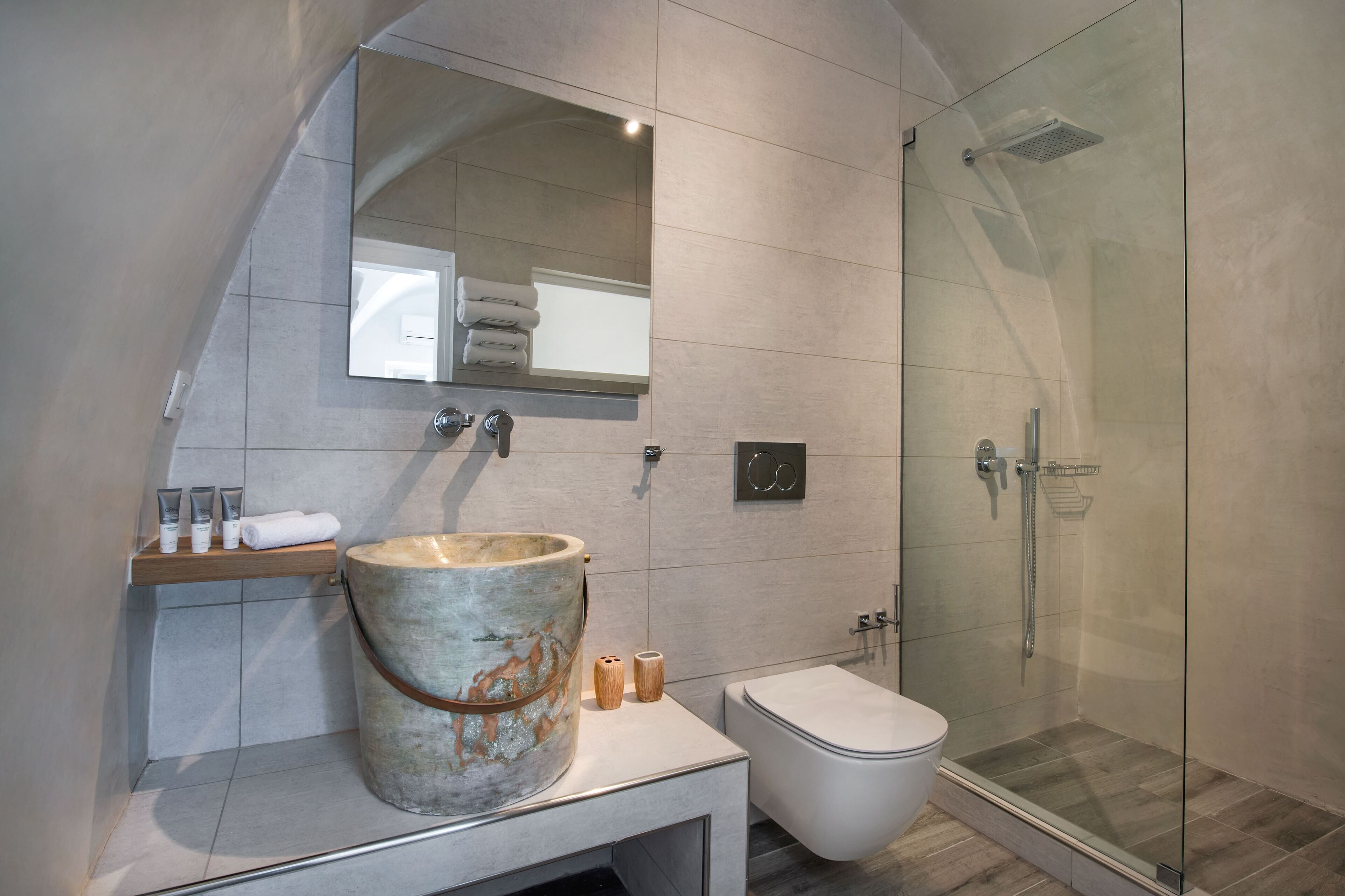 A modern bathroom with a large stone basin, a glass-enclosed shower, and a wooden shelf with toiletries.