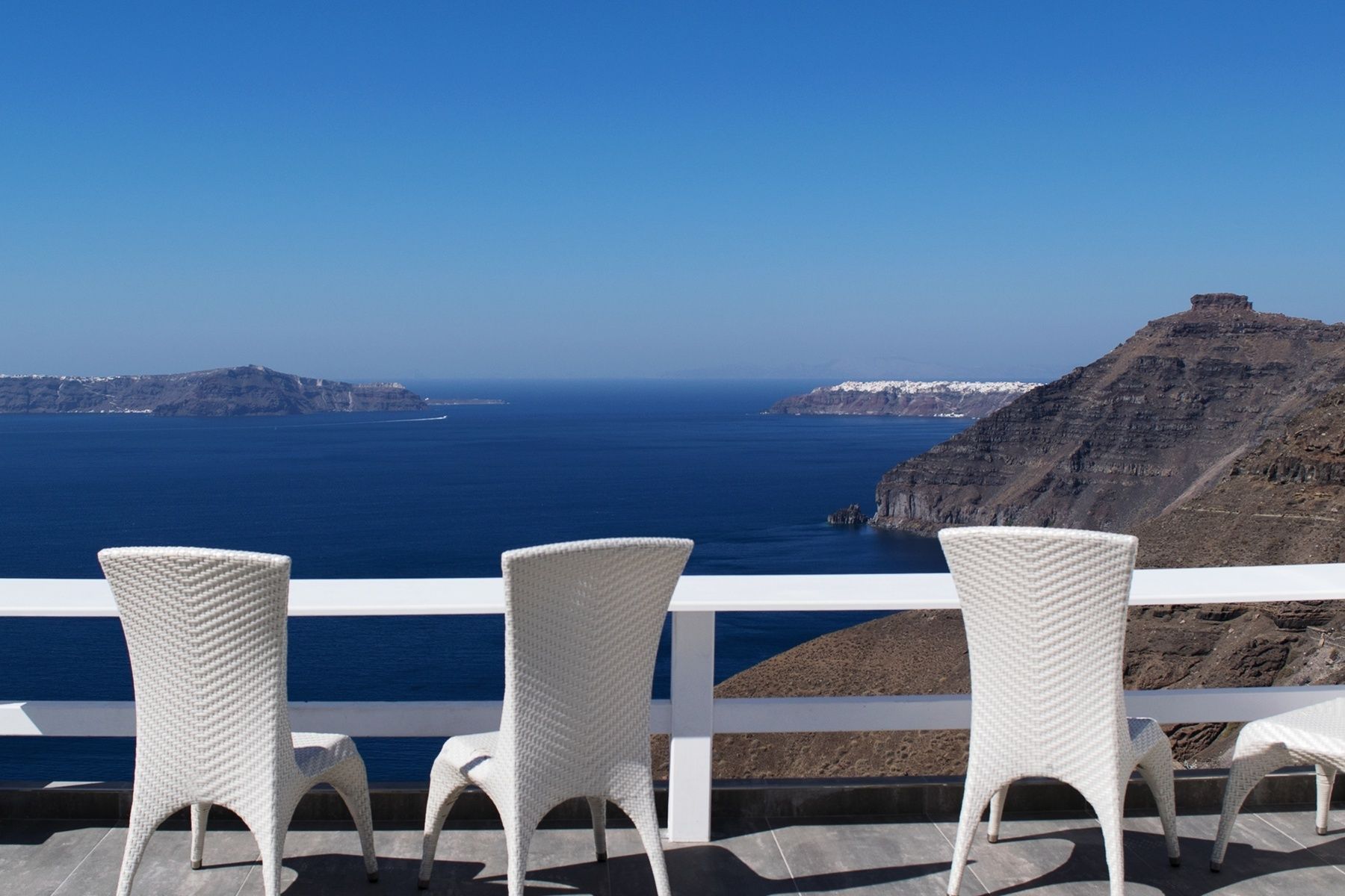 A balcony with white wicker chairs overlooking a coastal view with cliffs and the sea.