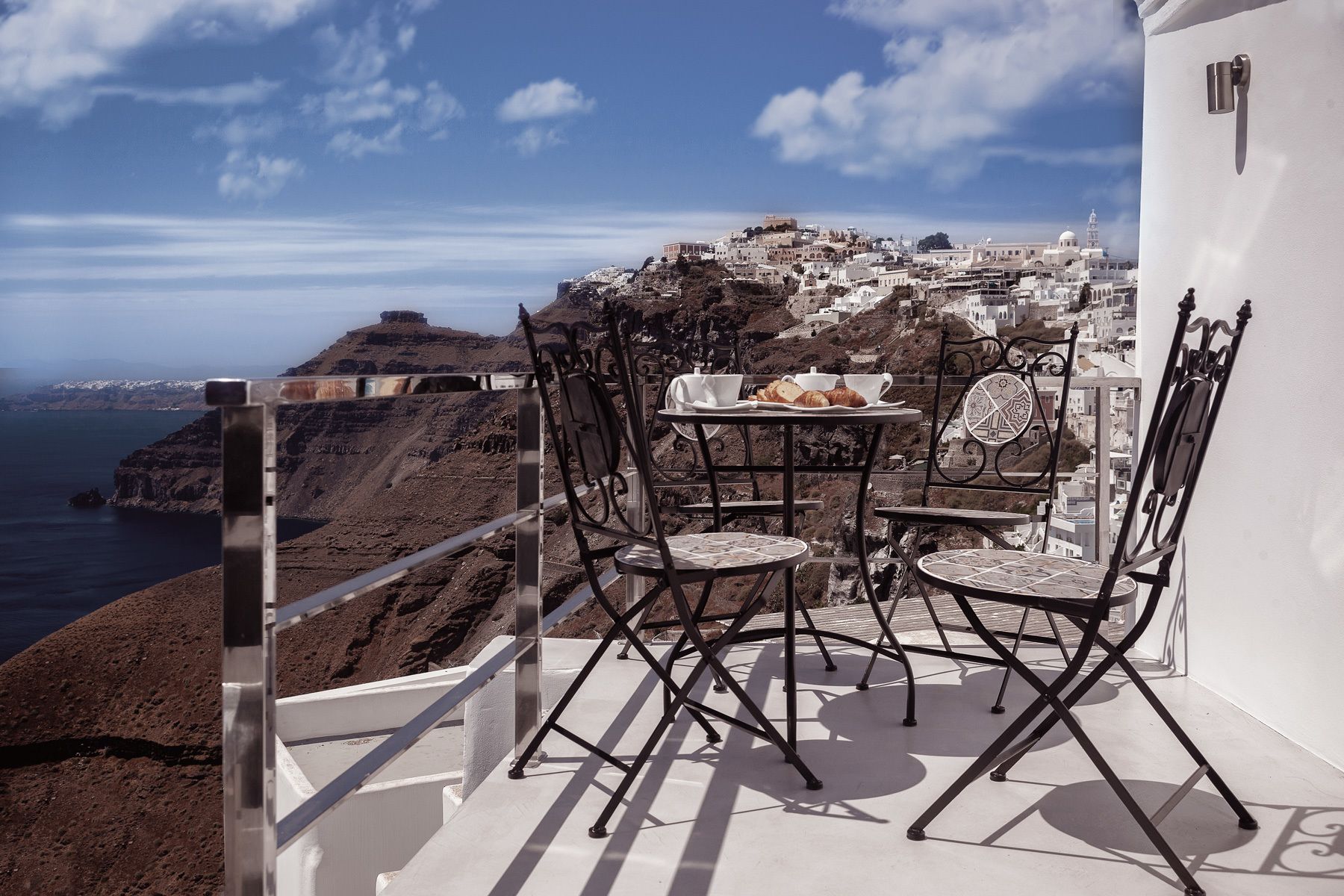 A balcony with a table and chairs overlooking a coastal town with white buildings and a view of the sea.