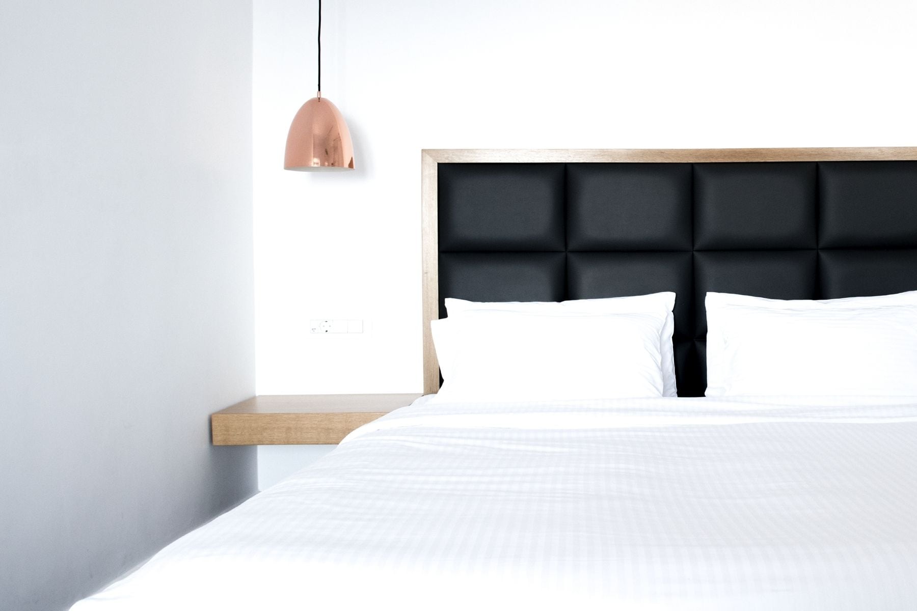 Un dormitorio moderno con una cama, una mesita de noche de madera, un cabecero tapizado negro y una lámpara colgante.