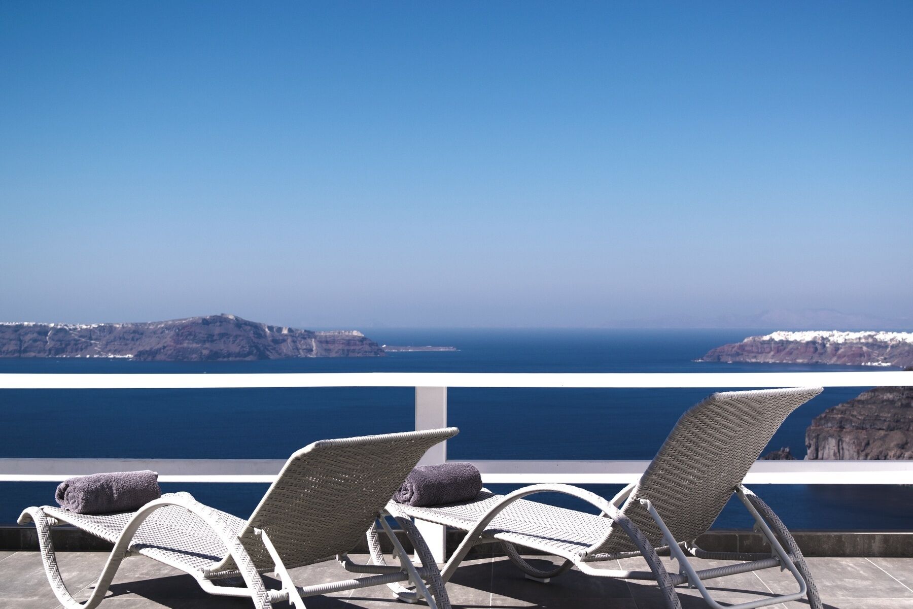 A balcony with two wicker lounge chairs, one with a towel, overlooking a coastal landscape with cliffs and the sea.