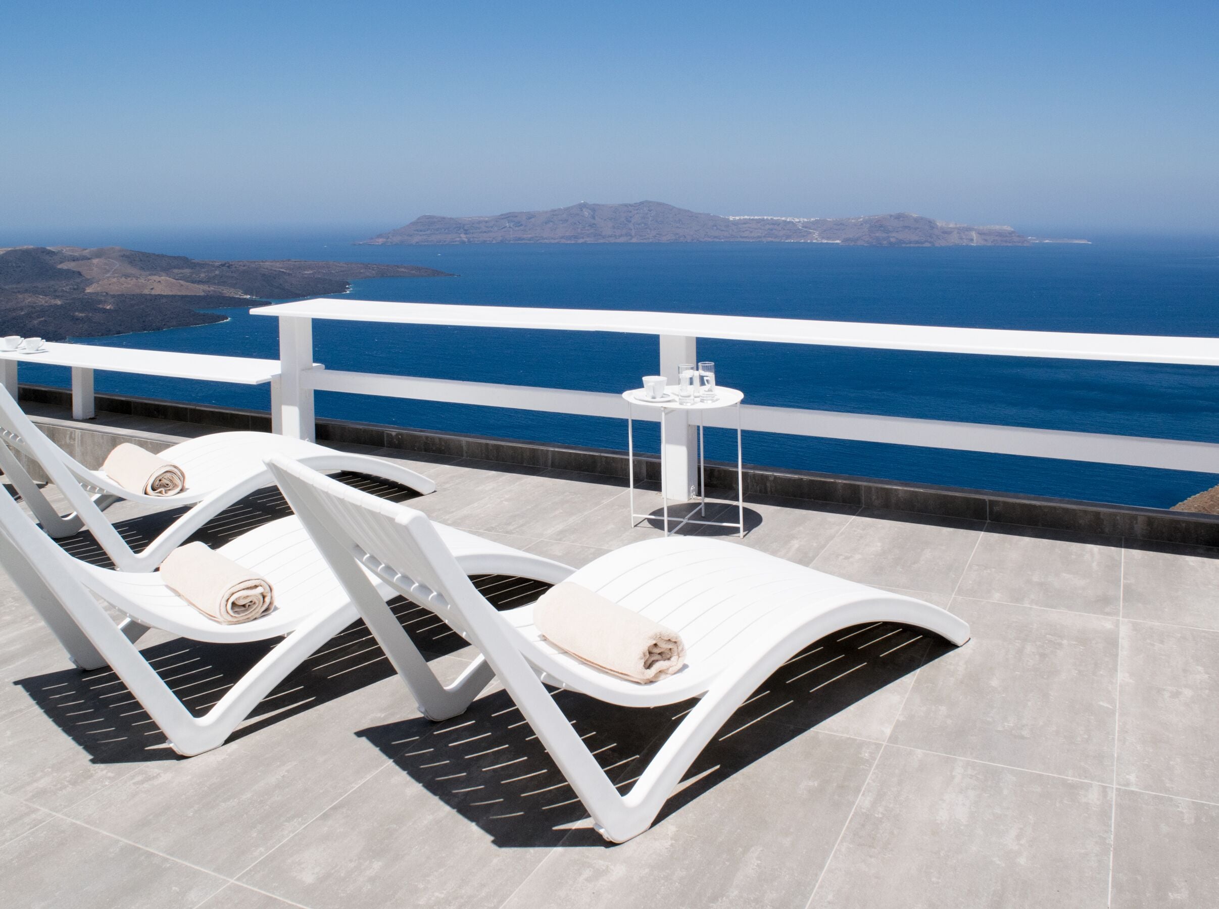 A terrace with white lounge chairs and a small table, overlooking a clear blue sea and distant land.