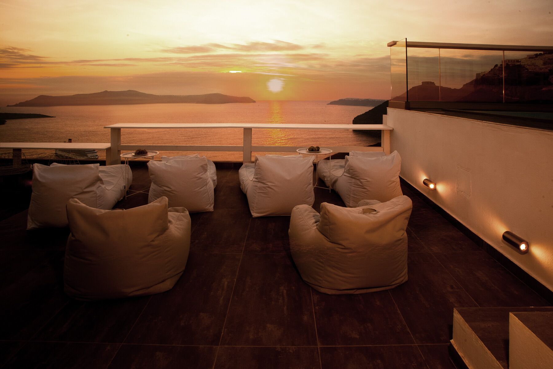 A sunset view from a balcony with seating and a glass railing overlooking the sea.