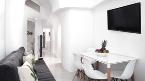 A modern, minimalist room with a white color scheme, featuring a sofa, a dining table with chairs, and a flat-screen TV mounted on the wall. - Thireas Hotel (Santorini)