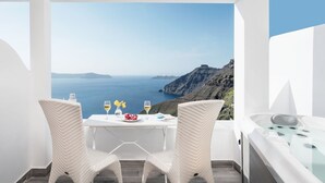 A balcony with a white table and two wicker chairs, overlooking a coastal landscape with cliffs and the sea. - Thireas Hotel (Santorini)