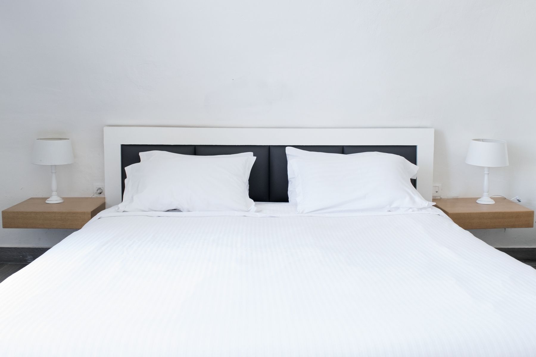 Una cama con ropa de cama y almohadas blancas, flanqueada por dos mesitas de noche con lámparas, frente a una pared lisa.