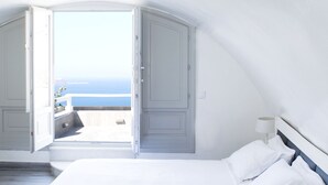 A bedroom with a bed, a nightstand, and a view of the sea through open doors. - Thireas Hotel (Santorini)