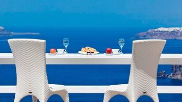 Una mesa con dos sillas de mimbre, un plato con pan, una manzana y dos vasos de agua, con vista al mar azul y el paisaje rocoso.