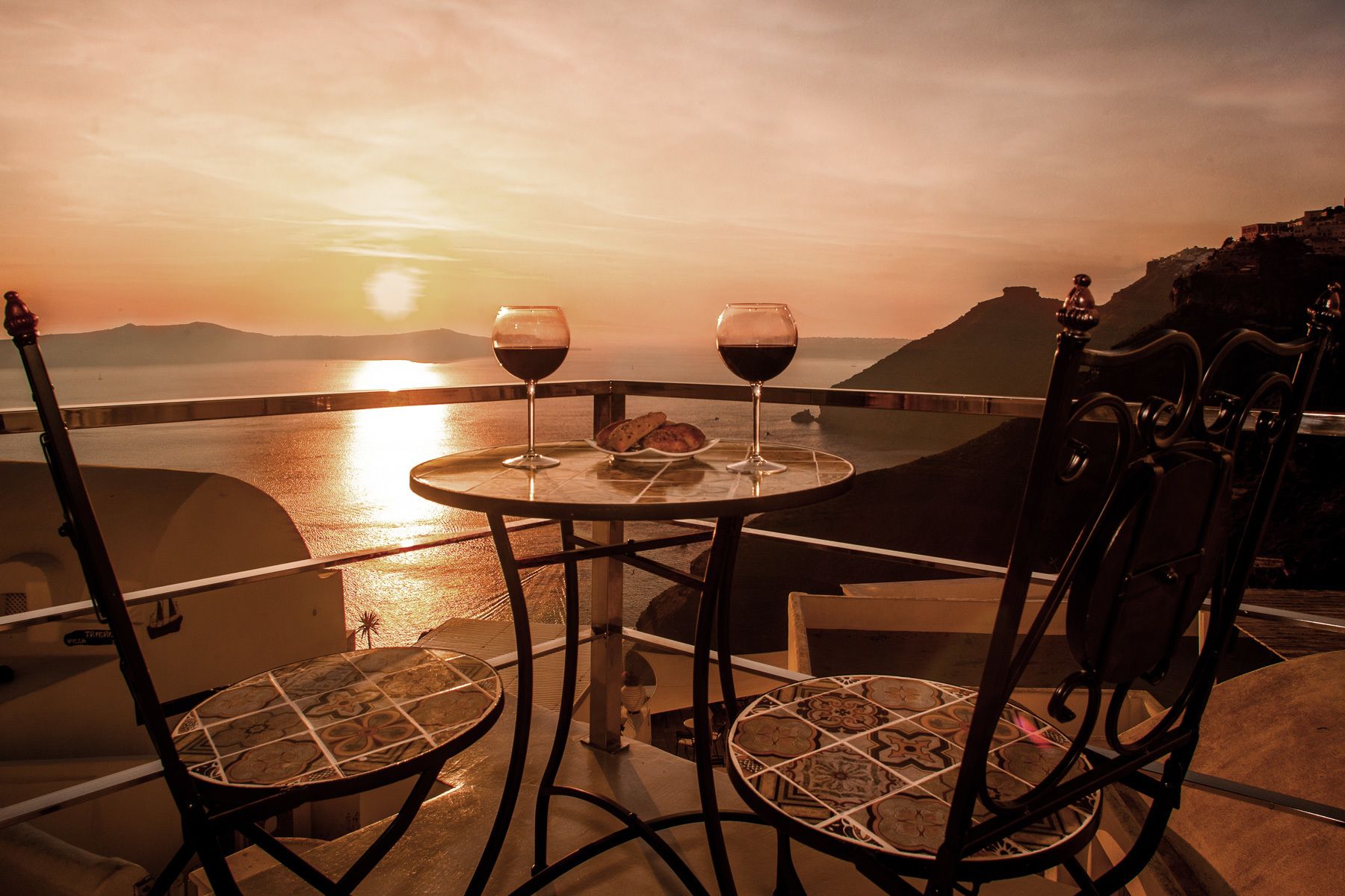 Une table avec deux verres à vin et une assiette de nourriture, placée sur un balcon donnant sur une étendue d’eau au coucher du soleil.