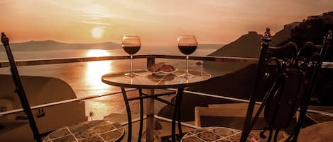 Une table avec deux verres à vin et une assiette de nourriture, placée sur un balcon donnant sur une étendue d’eau au coucher du soleil.
