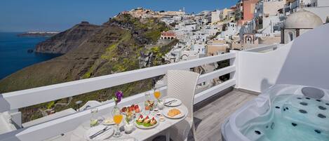 A balcony with a dining table set for breakfast, a hot tub, and a view of a coastal town.