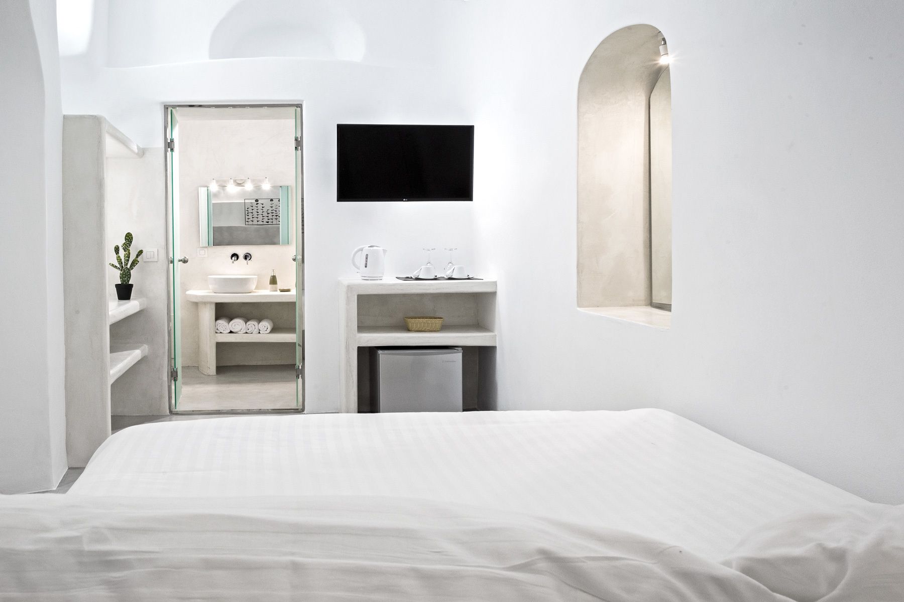 Habitación de hotel minimalista con una cama, un baño con lavamanos, un espejo y un televisor.